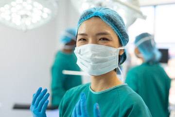 Smiling asian female surgeon in gown, mask, gloves and cap in operating theatre, copy space