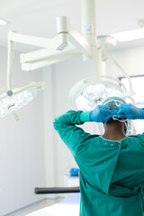 Rear view of african american surgeon in gown and gloves tying mask in theatre, vertical, copy space
