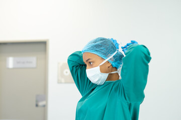 Biracial female surgeon in gown and cap tying on mask in operating theatre, copy space