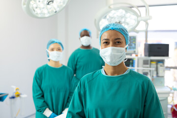 Fototapeta na wymiar Portrait of smiling biracial female surgeon in mask and cap in operating theatre with colleagues