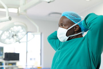 African american male surgeon in gown and cap tying mask in theatre, copy space