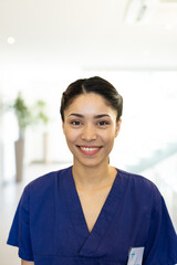 Vertical portrait of smiling biracial female healthcare worker in hospital corridor, with copy space
