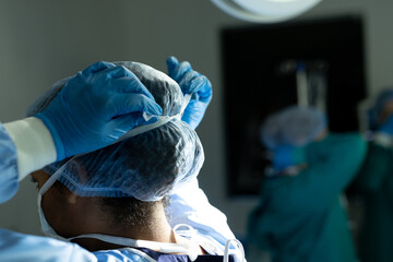 Rear view of biracial female surgeon in gloves and cap tying mask in theatre, with copy space - Powered by Adobe