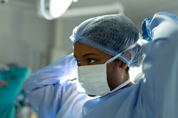 Biracial female surgeon in surgical cap and gown putting on mask in operating theatre
