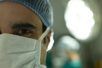 Fototapeta na wymiar Portrait of biracial male surgeon in surgical cap and mask in operating theatre with copy space