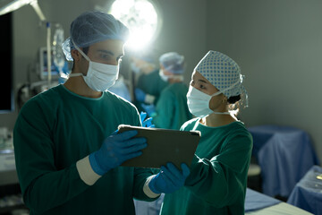 Diverse female and male surgeon in discussion, using tablet in operating theatre during surgery