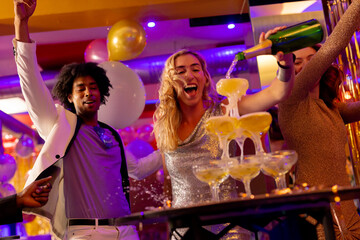 Happy caucasian woman pouring a champagne fountain into glasses with diverse friends at a nightclub