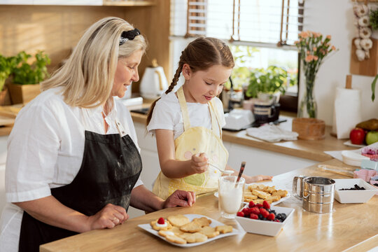 Happy delighted grandmother and grandchild baking cookies in kitchen standing at table Decorating cookies with sweet cream. Women preparing breakfast for mother. Young girl havin fun.