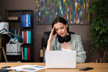 Worried woman with headache wearing modern earphones holding hand near face sitting at desk in office chair closing eyes. Tired lady after work study learning process from home.
