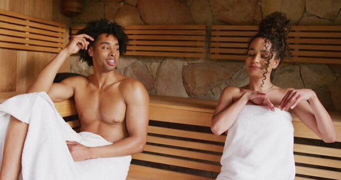 Video of relaxed happy diverse couple wearing towels sitting and talking in sauna room at health spa