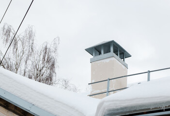 Smoke, square exhaust pipe on the roof of a modern house. The roof of the building is covered with white snow against the gray sky. Cloudy, cold winter day, soft light.