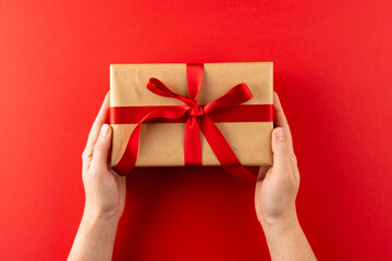 Overhead of hands holding gift in brown paper with red ribbon, on red background with copy space