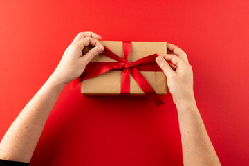 Overhead of hands tying gift in brown paper with red ribbon, on red background with copy space