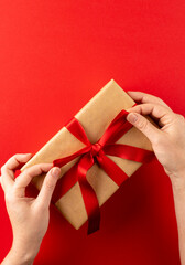 Vertical image of hands tying gift in brown paper with red ribbon, on red background with copy space
