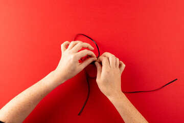 Obraz na płótnie Canvas Overhead of hands tying red heart gift box with black ribbon, on red background with copy space