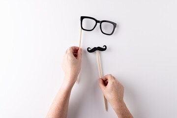 Hands holding black false moustache and glasses on sticks, on white background with copy space