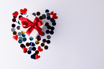 White gift box with red ribbon on heart shape of confetti, on white background with copy space