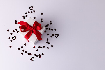 White gift box with red ribbon on heart confetti, on white background with copy space