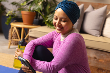 Happy biracial woman wearing hijab, sitting on yoga mat and using smartphone in living room