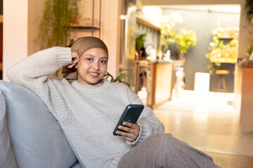 Portrait of happy biracial woman wearing hijab, sitting on sofa and using smartphone