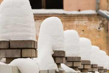 High, even, columns of snow on the brick pillars of the fence. An even row of small snowdrifts against the background of a shabby wall in defocus. Urban winter landscape. Cloudy winter day, soft light