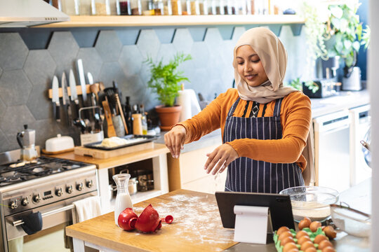 Happy biracial woman wearing hijab, standing in kitchen and baking