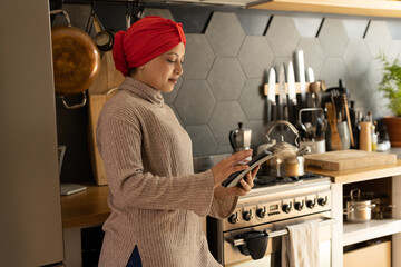 Happy biracial woman wearing hijab, standing in kitchen and using smartphone