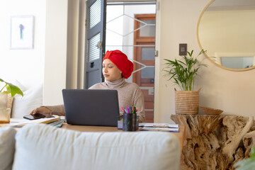 Happy biracial woman wearing hijab, sitting at table, using laptop and smartphone