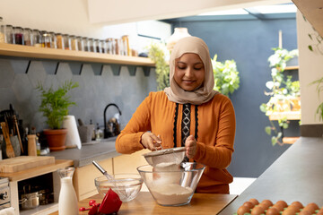 Happy biracial woman wearing hijab, standing in kitchen and baking