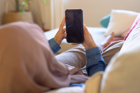 Happy biracial woman wearing hijab, sitting on sofa in living room, using smarphone
