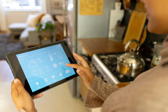 Happy biracial woman wearing hijab, standing in kitchen and using tablet with weather forecast