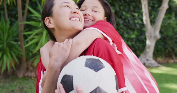 Video of happy asian mother and daughter in soccer shirts embracing in garden