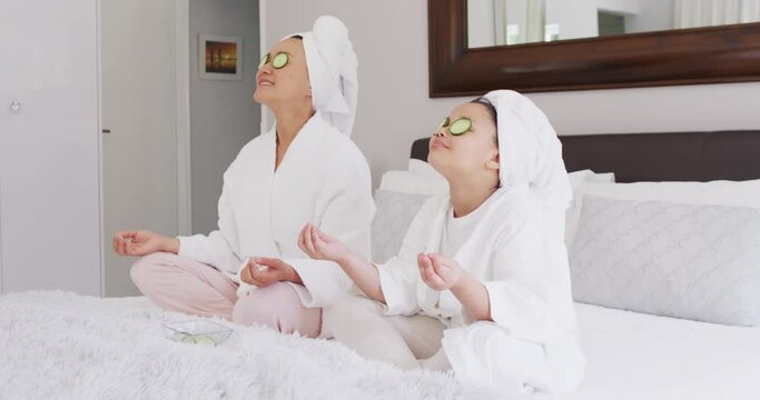 Video of happy asian mother and daughter in robes doing cucumber mask and resting
