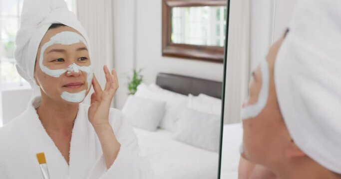 Video of happy asian woman in robe with moisturizing mask looking at mirror