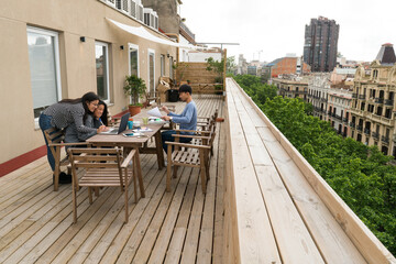 Side view of three young people sitting at table on balcony and working with gadgets.