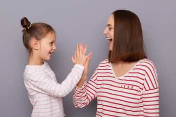 Image of excited extremely happy mother and little daughter wearing casual style shirts, making high five gesture, having done all things perfectly, posing isolated over gray background.