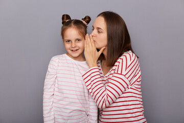 Brown haired woman and little girl with two hair buns posing isolated over gray background, mother whispering secret to her kid, covering her ear with palm, nobody hearing private information.