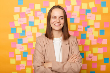 Indoor shot of confident businesswoman wearing beige jacket standing against yellow wall covered...