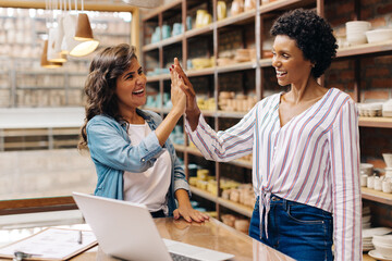 Successful female ceramists high fiving each other in their shop. Women owning a small business in...