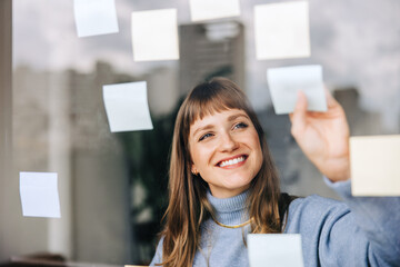 Cheerful young businesswoman sticking adhesive notes to a glass wall
