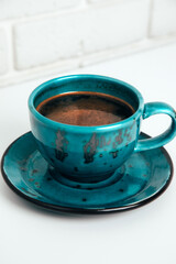 Blue mug with black coffee with a saucer on a white table. Front view, close-up