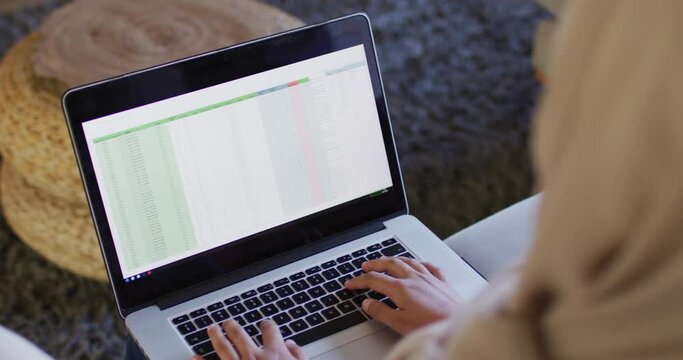 Over shoulder video of biracial woman in hijab at home working on laptop, spreadsheet on screen