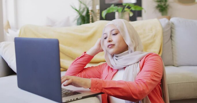 Video of concentrating biracial woman in hijab at home sitting on floor working on laptop