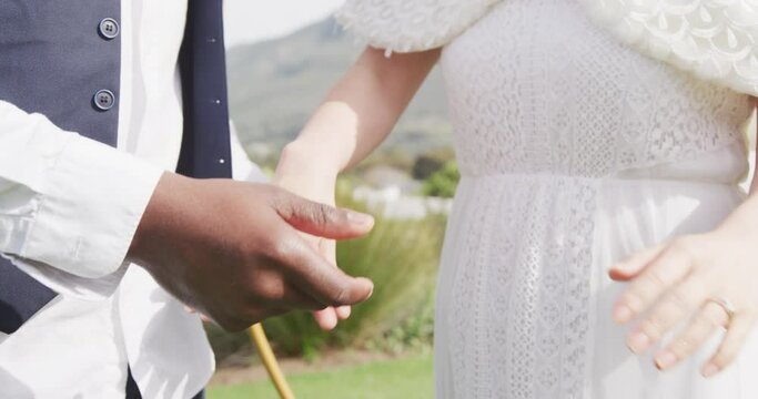 Video of midsection of diverse bride and groom holding hands and walking at outdoor wedding
