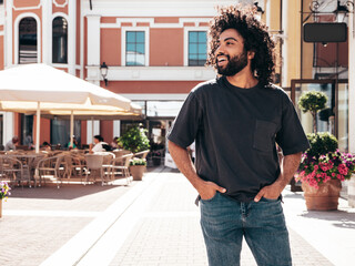 Fototapeta premium Handsome smiling hipster model.Sexy unshaven Arabian man dressed in summer clothes. Fashion male with long curly hairstyle posing in the street at sunset. Cheerful and happy