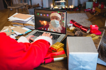 Santa claus with christmas presents having video call with caucasian santa claus with face mask