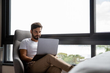 Young man sitting in front of his laptop, chatting, shopping, successful freelancer working online on computer, sitting in an armchair in his bedroom 