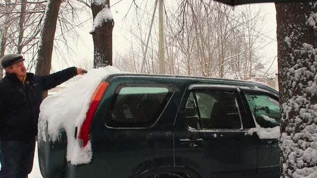 A man cleans the rear window of the car from snow with a brush, it is snowing. The car is covered with snow after a snowstorm and looks like a snowdrift. City life, heavy snowfall. UHD 4K.