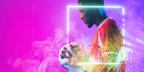 African american male soccer player with ball standing by illuminated plant and square