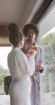Vertical video of diverse couple in bathrobes drinking tea and talking by window at health spa